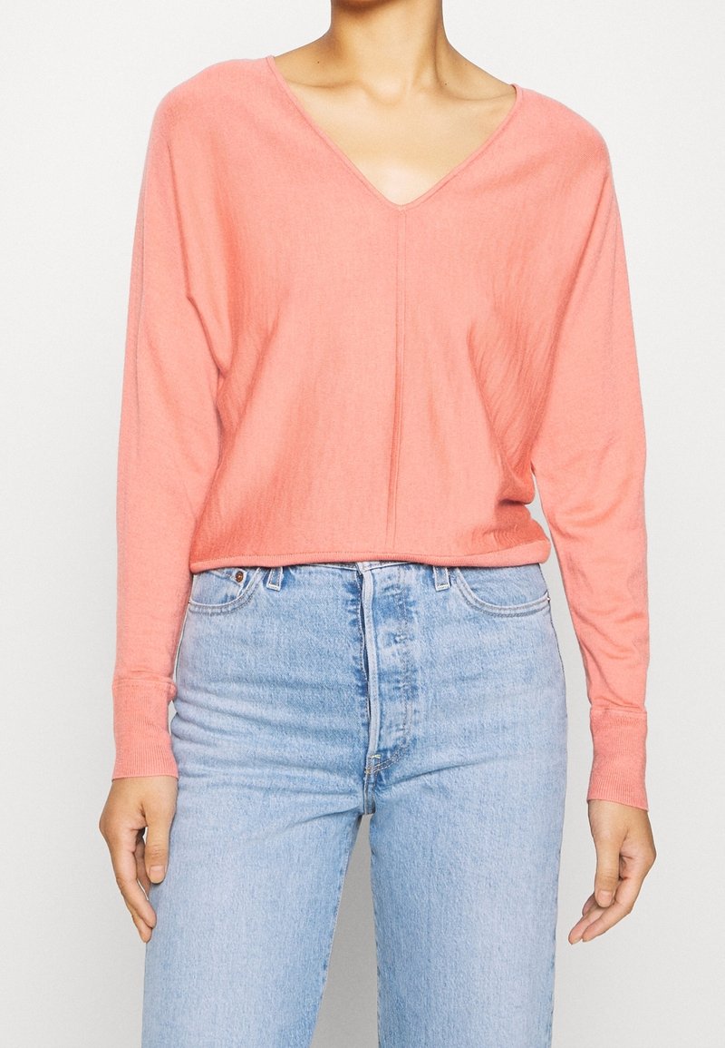 Korallenfarbenes, oversized Langarmshirt mit tiefem V-Ausschnitt, aus strukturiertem Stoff. Kombiniert mit hoch geschnittenen hellblauen Jeans.