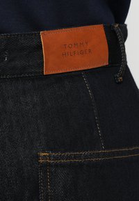 Sötétkék farmernadrág barna bőrcímmel, amelyen a "TOMMY HILFIGER" felirat domborodik ki, látható varrási részletekkel és klasszikus öt zsebes kialakítással.