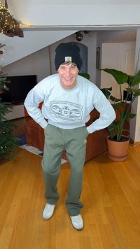 Grauer Sweatshirt mit grafischem Aufdruck, grüne Jogginghose und weiße Sneaker. Schwarze Mütze und Innenraum mit Pflanze und Sofa.