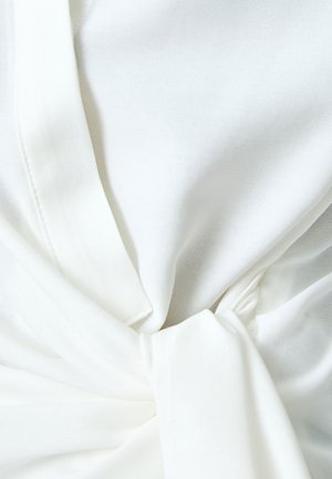 Top de tela blanca con un detalle de nudo en la cintura, que presenta una textura suave y un diseño ligero y drapeado. Las costuras son visibles.