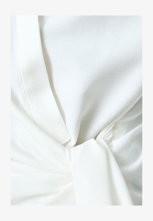 Weißes Oberteil aus Stoff mit einem Knoten-Detail an der Taille, das eine glatte Textur und ein leichtes, drapiertes Design aufweist. Die Nahtnähte sind sichtbar.