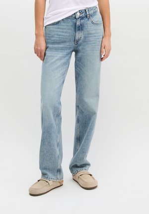 Osoba ubrana w jasnoniebieskie jeansy o prostych nogawkach, biały top i beżowe mokasyny wsuwane, stojąca na tle jednolitego jasnego tła.