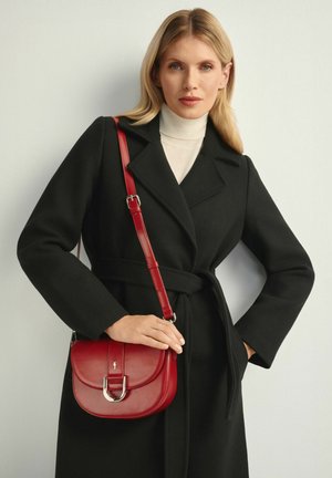 Kobieta w czarnym płaszczu z paskiem i białym golfem, trzymająca czerwoną skórzaną torebkę na ramię z kozłami metalowymi.
