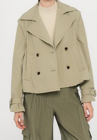Veste croisée kaki avec grands revers, poches avant et accents à boucle sur les manches, associée à un haut beige clair et un pantalon assorti.