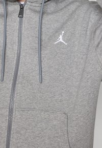 Sudadera con capucha gris claro de cremallera, hecha de una tela suave, que presenta un bolsillo tipo canguro, cordones grises y un logo Jumpman blanco en el pecho.