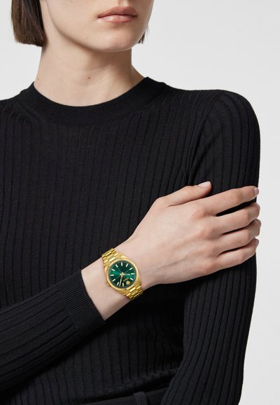 Zeltīts rokas pulkstenis ar zaļu ciparnīcu, kas aprīkots ar baltām stundu atzīmēm un teksturētu zeltītu siksnu. Valkāts uz rokas pret melnu ribotu džemperi.
