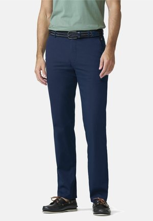 Man draagt marineblauwe broek, zwarte gevlochten riem, lichtgroen overhemd en zwarte bootschoenen met bruine veters, staand op een witte achtergrond.