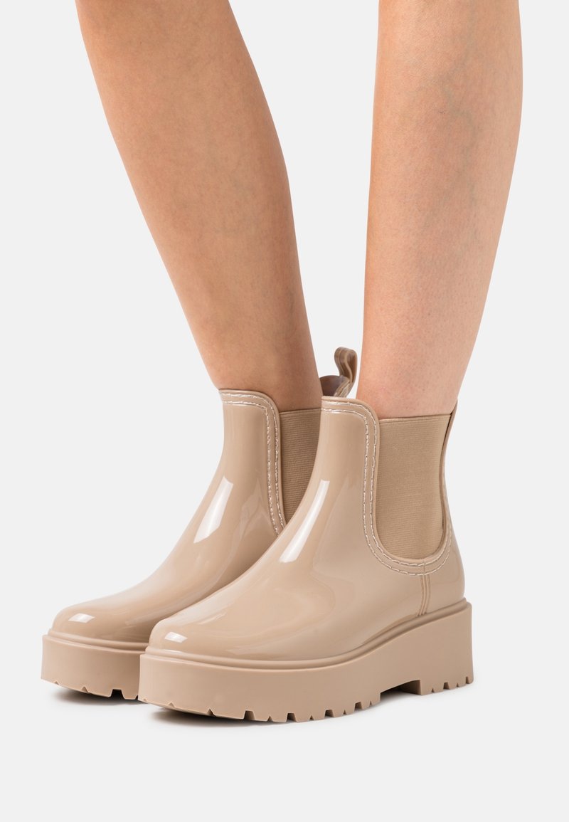 Botas de tobillo de goma en color beige con un acabado brillante, paneles laterales elásticos y suela gruesa, que presentan detalles de costura en contraste.