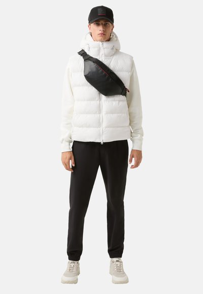 Witte puffer vest met hoge kraag, zwarte schoudertas, zwarte broek en neutrale sneakers. Eenvoudig ontwerp met functionele details.