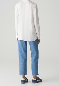 Blouse blanche à manches longues associée à un jean denim bleu clair et des baskets bleu marine avec trois bandes blanches sur le côté.