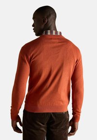 Oranžový pletený sveter s rebrovanými rukávmi a lemom, s prúžkovaným golierom viditeľným zozadu, nosený cez tmavé kordové nohavice.