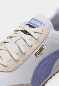 Κοντινό πλάνο λευκού αθλητικού παπουτσιού Puma Arizona με κρεμ κορδόνια, μπεζ σουέτ λεπτομέρειες και μωβ ρίγα στο πλάι που δείχνει το χρυσό λογότυπο Puma.