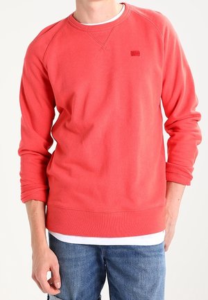 Mand iført en koralrød crewneck-sweatshirt over en hvid skjorte med blå jeans, stående foran en ensfarvet baggrund.