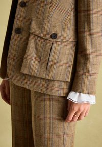 Hnedý károvaný oblek s dvojradovým saka s dvoma gombíkmi a našitým vreckom, v kombinácii so zodpovedajúcimi nohavicami a bielym detailom na rukávoch.