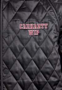 Quiltet sort jakke med en diamantmønster, med trykknapper. Broderet "Carhartt WIP" i rød foran. Glat tekstur.