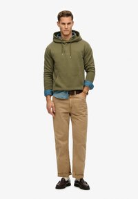 Olivgrön hoodie med en framficka, lagd över en ljusblå denimskjorta, beige chinos och svarta loafers. Enkel, avslappnad stil.