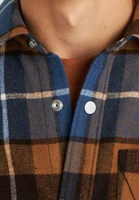 Koszula flanelowa w kratę w kolorach niebieskim, brązowym i kremowym. Posiada zatrzaski i kieszeń na piersi, z miękką teksturą i ciepłym wyglądem.