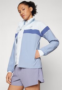 Giacca a vento blu chiaro con strisce blu scuro, chiusura con zip e polsini elasticizzati, abbinata a pantaloni corti color lavanda.