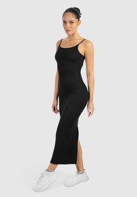 Donna in piedi con un vestito midi nero senza maniche e sneakers bianche su uno sfondo grigio chiaro.