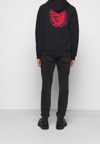 Sudadera negra con un gráfico rojo en la parte trasera, que presenta una cara sonriente y texto. Combinada con jeans negros y botas negras. Diseño simple y casual.