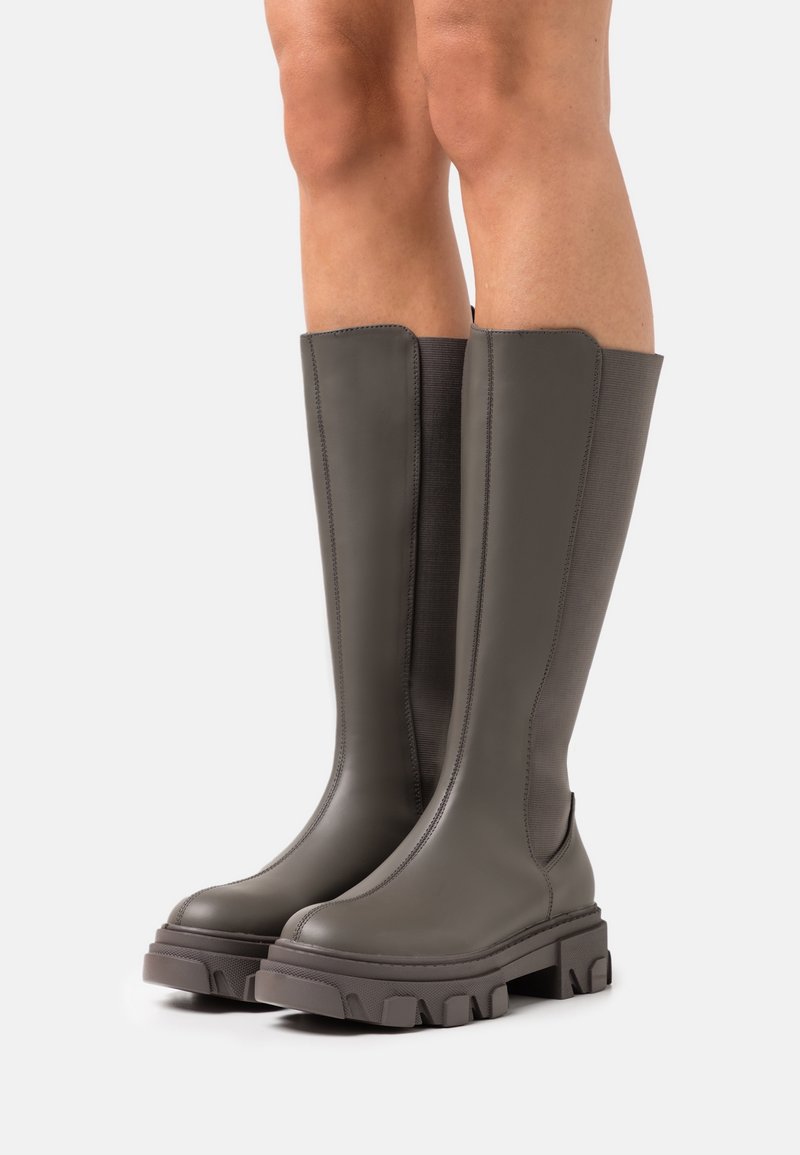 Botas hasta la rodilla en gris oscuro, con un acabado similar al cuero suave, suela gruesa con tracción y paneles laterales elásticos para ponerlas y quitarlas fácilmente.