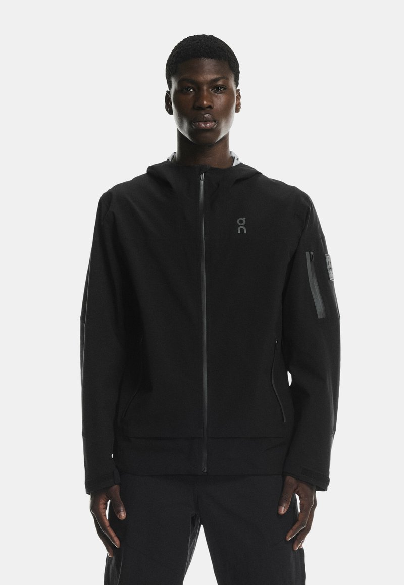 Černá kapucínová bunda se zipovým zapínáním, vyrobená z hladkého materiálu. Obsahuje logo na hrudi a zipovou kapsu na rukávu.