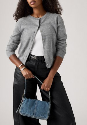 Frau in grauem Cardigan, weißem Rippenshirt, schwarzen Hosen mit Gürtel, die eine Jeanshandtasche an dem Griff vor einem neutralen Hintergrund hält.