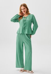 Completo verde a due pezzi con texture, composto da un top a peplo con dettagli a fiocco e pantaloni a gamba larga, realizzato in tessuto morbido. Sandali chiari e neutri.