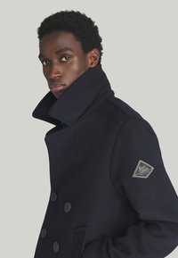 Cappotto di lana navy con colletto alto e design doppiopetto. Presenta grandi bottoni e una toppa con logo sulla manica sinistra.