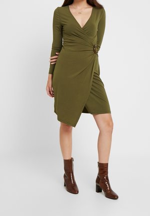 Woman wearing olive green wrap dress with long sleeves and brown ankle boots, standing against a plain light background.