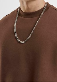 Felpa marrone con un colletto a coste, sovrapposta ad una collana d'argento con un design a catena a maglie larghe.