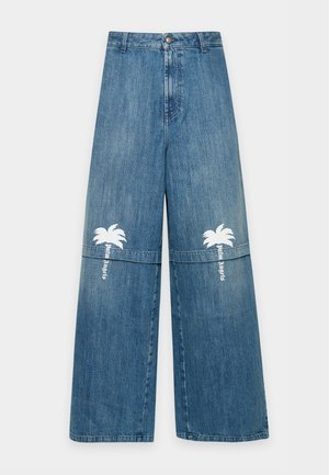 Un homme pose devant un fond uni, vêtu d’une casquette et d’un tee-shirt noirs, d’un jean bleu orné de motifs de palmiers blancs, de baskets noires et d’un bracelet argenté.