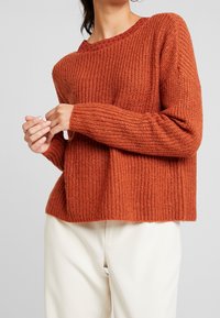 Femme portant un pull en maille grossière de couleur rouille et un pantalon blanc, ajustant sa manche avec les deux mains devant un fond gris.