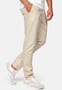 Bež chinos hlače sa slim fit krojem, ravnim nogavicama, bočnim džepovima i suptilnom teksturom. Uparene s bijelim i bež sportskim tenisicama.