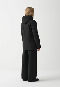 Zwarte hoodie met een gladde textuur, met een relaxte pasvorm en schuine naden. Gecombineerd met zwarte wijde broek en platte schoenen.