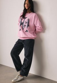 Roze oversized sweatshirt met visafbeelding, gecombineerd met donkere wijde broekspijpen en zilveren sneakers. Het materiaal lijkt zacht en comfortabel.