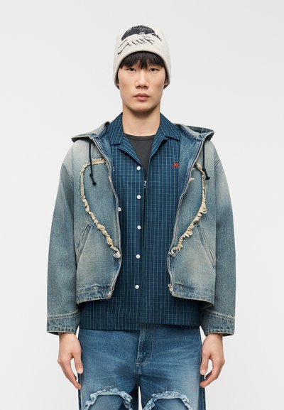 Νεαρός άνδρας που φοράει σκισμένο τζιν μπουφάν, καρό πουκάμισο, σκισμένο τζιν και μπεζ σκουφάκι με μαύρο γραφικό σχεδιασμό.