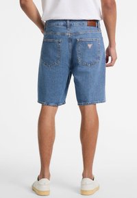 Pantalones cortos de denim en azul claro, con corte recto, diseño de múltiples bolsillos y una etiqueta de cuero en la parte trasera de la cintura. Zapatos deportivos blancos en los pies.