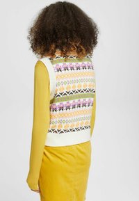 Gilet en tricot de couleur crème avec des motifs géométriques multicolores, porté sur un top jaune moutarde. Encolure ronde et détail de bord côtes.