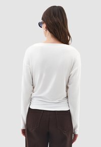 Bílý top s dlouhým rukávem a volným střihem, s překládaným zadním dílem. Doplněno tmavě hnědými kalhotami s dvěma zadními kapsami a viditelným švem.