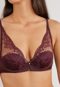 Soutien-gorge en dentelle bourgogne foncé avec des motifs floraux, un décolleté plongeant et un petit détail décoratif au centre. Panneaux en mesh doux inclus.