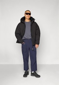 Črna puhovka s kapuco, siva majica z kratkimi rokavi, mornarsko modre cargo hlače in črni supergi. Ponaša se z žepi in sproščenim krojem.