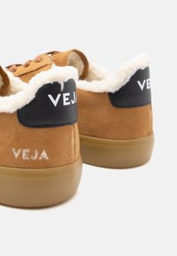 Smeđe nubuk tenisice s crnim kožnim detaljima, pahuljastim bijelim podlogama i gumiranim potplatima. Imaju urezano "VEJA" logo na peti i sa strane.