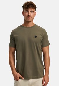 Olivengrünes Baumwoll-T-Shirt mit kurzen Ärmeln, Rundhalsausschnitt und einem kleinen schwarzen runden Logo auf der linken Brust. Glatte Textur, lässige Passform.
