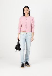 Ανοιχτό ροζ πλεκτό μπουφάν με στρογγυλή λαιμόκοψη και κουμπιά, συνδυασμένο με ανοιχτό μπλε τζιν και μαύρη τσάντα με σχέδιο. Περιλαμβάνονται μαύρα αθλητικά παπούτσια.