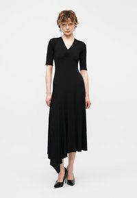 Γυναίκα που φοράει μαύρο ασύμμετρο μίντι φόρεμα με κοντά μανίκια, λαιμόκοψη σε σχήμα V και μαύρα παπούτσια με ψηλά τακούνια, στέκεται μπροστά σε απλό λευκό φόντο.