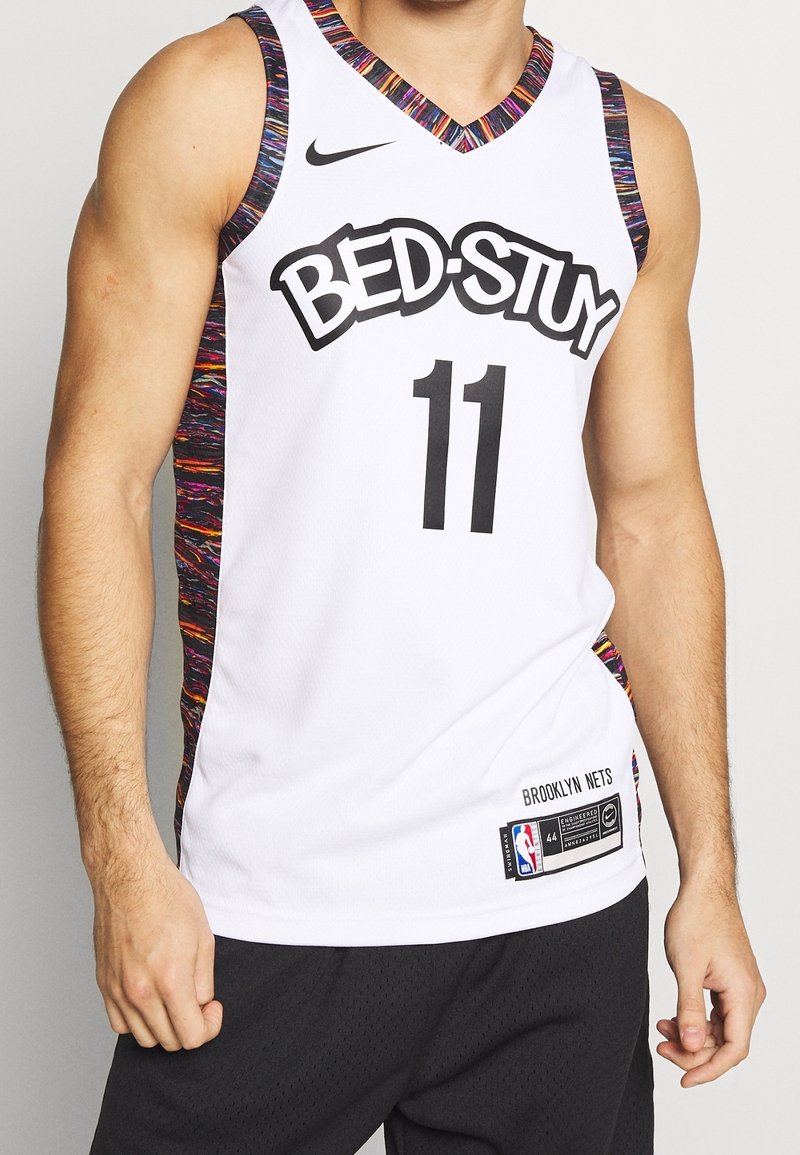 Maillot de basket-ball blanc sans manches avec une bordure colorée, texte "BED-STUY", numéro 11 et logo Brooklyn Nets NBA porté par une personne.