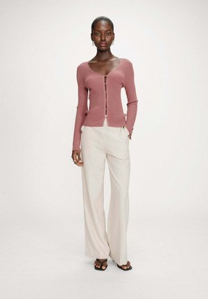 Femme portant un cardigan boutonné rose poussiéreux, un pantalon large crème, des sandales noires à bout carré et des petites boucles d'oreilles en anneau, sur un fond blanc.