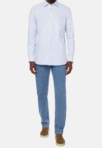 Mužský model v světle modré a bílé pruhované košili s knoflíky, modrých džínách a hnědých teniskách, pózující na jednobarevn ém pozadí.