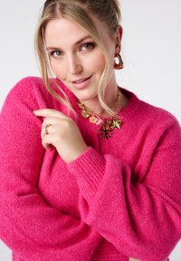 Roze gebreide trui met brede mouwen en een gestructureerde afwerking. Gouden bloemenketting met een ketting en roze accenten.